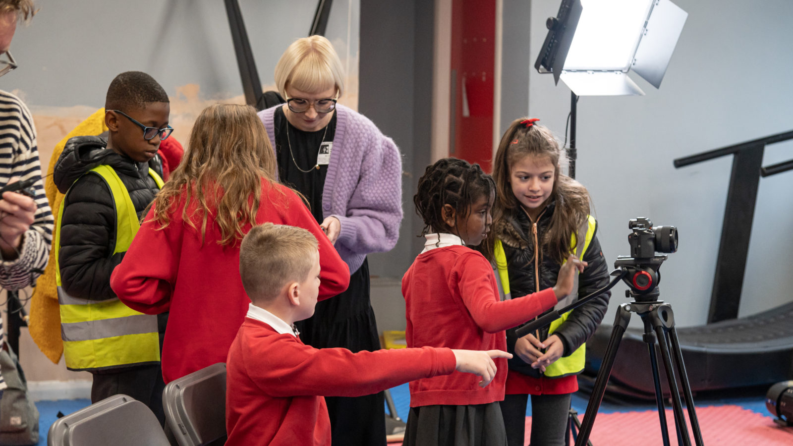 Photograph of a group of children in red school uniforms, using a camera and other recording equipment. They are being helped by a woman with a blonde bob, glasses and lilac cardigan