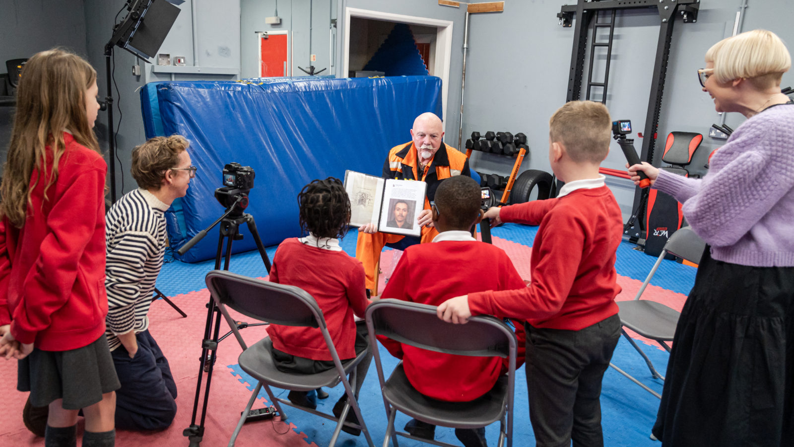 Photograph of a group of children in red school uniforms, seated around a man wearing an orange hi-vis vest, The children are interviewing him and filming with a go pro camera