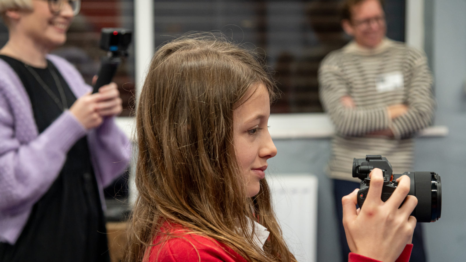 Photograph of a schoolgirl holding a camera, aimed at someone out of shot. In the background a woman with a blonde bob, glasses and lilac cardigan smiles