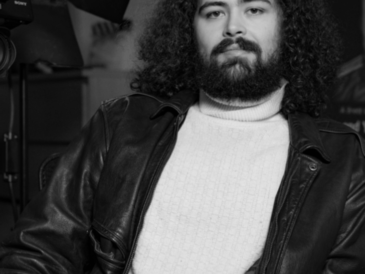 Photograph of a white man with dark, curly hair wearing a white turtleneck jumper, sitting in front of a studio lamp