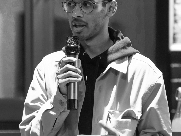 Photograph of a dark skinned man in a baseball cap and glasses. He is speaking into a microphone