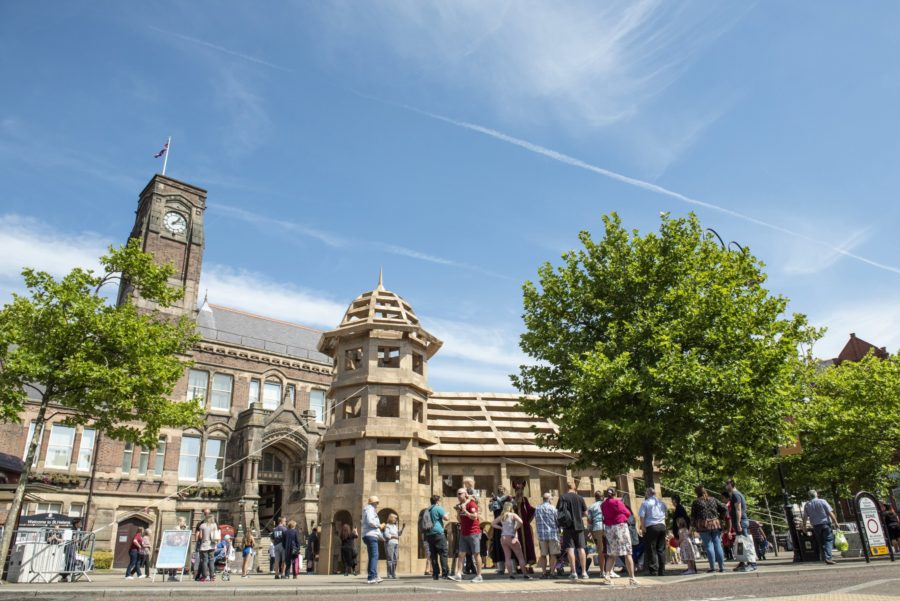 A crowd of people are gathered underneath a huge castle made of cardboard. It is a sunny warm day with blue skies. The castle sits in front of a large Town Hall building.