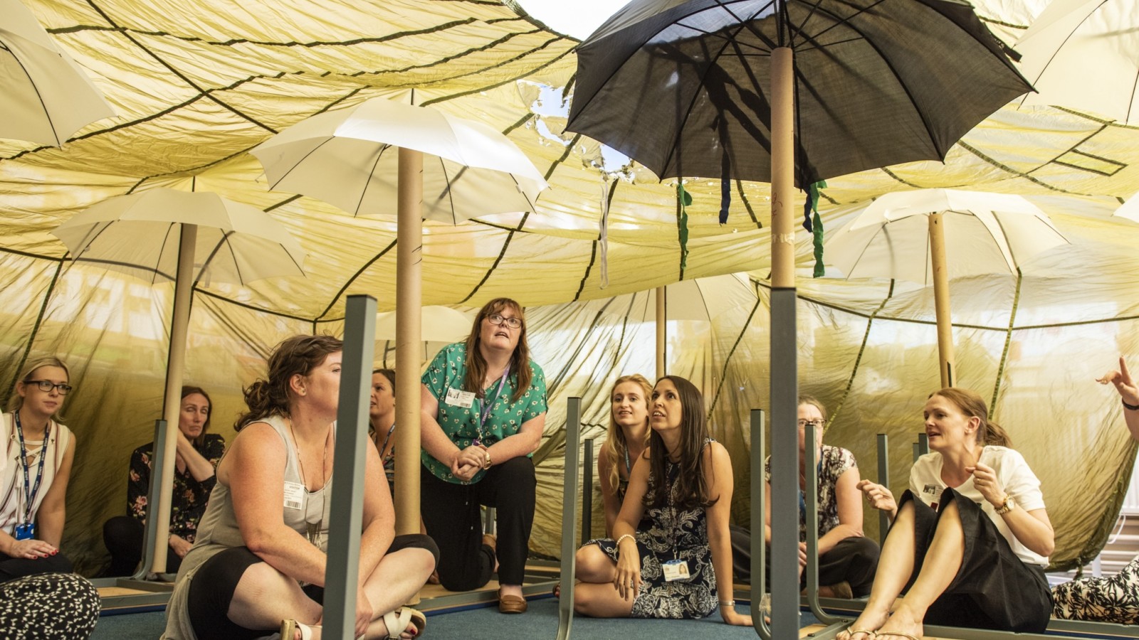5 women sit in a bell tent with umbrellas hanging from the ceiling. The women are listening to another woman who is talking to the group. There is an upturned table in front of the woman speaking, who has dark hair and is wearing a cream coloured top.|